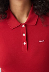 Červené polo tričko vyrobené z texturované tkaniny, které má límeček, tři bílé knoflíky a malé logo Levi's na levém prsu.