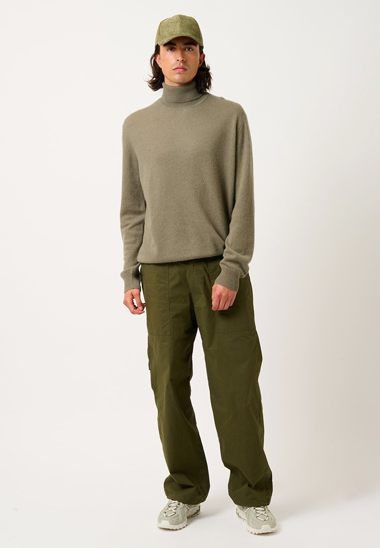 Mann steht mit einem olivgrünen Rollkragenpullover, passendem Hut, lockeren olivfarbenen Cargo-Hosen und hellgrauen Sneakers vor einem schlichten Hintergrund.