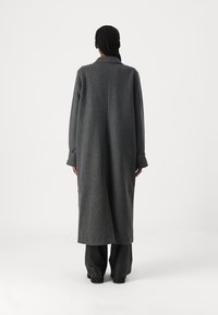 Cappotto lungo in lana grigia con colletto, vestibilità over, maniche larghe e silhouette pulita, abbinato a pantaloni neri su uno sfondo neutro.