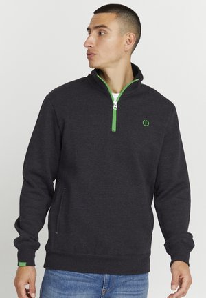 Sudadera de cremallera color gris oscuro con acentos verdes, que presenta un cuello alto, bolsillos laterales y un logo en el pecho. Tejido texturizado.
