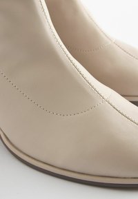 Stivaletti in pelle beige con texture liscia, punte appuntite e cuciture a contrasto. Presentano un tacco basso e affusolato e un design elegante.