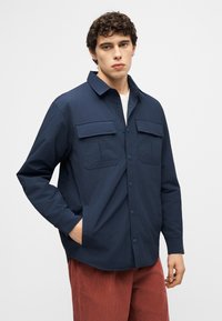 Jeune homme portant une chemise à boutons bleu marine avec des poches poitrine et un pantalon en velours côtelé couleur rouille, debout avec une main dans la poche.