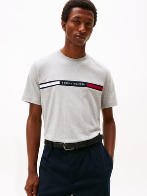 Szary bawełniany t-shirt z poziomym pasem z logo "TOMMY HILFIGER" w granatowym kolorze, podkreślony czerwonymi i białymi paskami. Noszony z granatowymi spodniami.