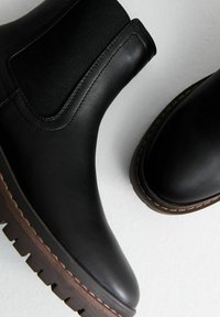 Botas de cuero negro hasta el tobillo con paneles elásticos, puntera redondeada y suelas texturizadas en marrón. Costuras visibles que destacan a lo largo de los bordes.