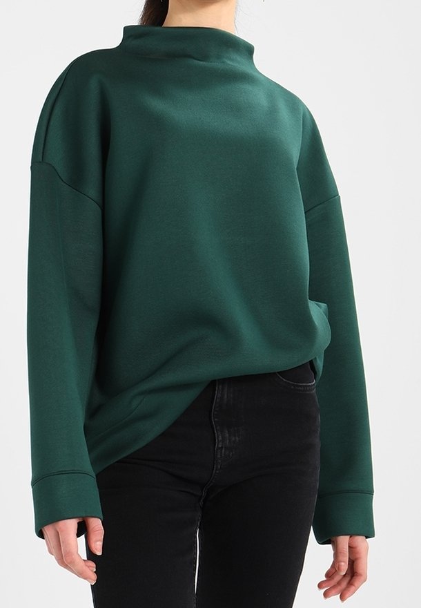 Vrouw in een donkergroene oversized sweatshirt met lange mouwen en een ontspannen hoge halslijn, gecombineerd met zwarte aansluitende broek.