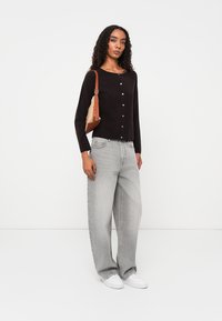 Cardigan preto de mangas longas com decote arredondado e fecho de botão, combinado com jeans cinzentos largos e ténis brancos. Acessório em forma de bolsa castanha.