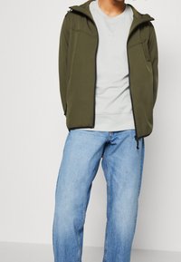 Oliivinvihreä softshell-takki hupulla ja mustalla vetoketjulla; päällä vaaleanharmaa collegepaita ja yhdistetty vaaleansinisiin farkkuihin.