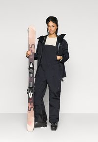 Μαύρο μπουφάν σκι με προσαρτημένη κουκούλα, μαύρες φόρμες, κρατώντας ροζ και μαύρα σκι. Υφή υλικού με φερμουάρ, μποτάκια σκι και γυαλιά.