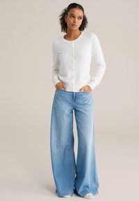 Cardigan alb texturat cu închidere cu nasturi, asortat cu blugi сu croială largă într-o nuanță deschisă de albastru. Modelul pozează cu mâinile în buzunare, pe un fundal neutru.