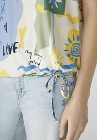 Blusa colorata con motivi astratti in blu, verde e giallo. Presenta un orlo regolabile con coulisse e una superficie in tessuto liscia.