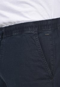 Pantalons bleu marine à taille élastique avec un motif texturé et côtelé. Comprend une poche latérale et une petite étiquette de marque à la taille.