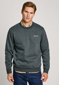 Sudadera gris con puños y dobladillo de canalé, con un pequeño logo blanco. Lleva puesta sobre una camisa de cuadros, exhibida contra un fondo neutro.
