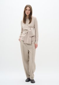 Beige langärmliges Oberteil mit Knoten-Detail, kombiniert mit lockeren beigen Hosen und schwarzen Slippers vor einem schlichten Hintergrund.
