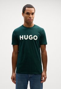 T-shirt de algodão verde com mangas curtas, decote redondo e grande texto branco "HUGO" no peito. Corte casual e textura suave.