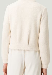 Cardigan lavorato a maglia di colore crema con design corto e polsini a coste, caratterizzato da una semplice scollatura tonda e senza chiusure visibili.