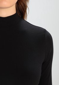 Sort langærmet top med høj hals, lavet af glat, strækbart stof. Har en tætsiddende silhuet og sømløst design.