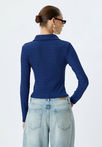 Maglietta a maniche lunghe blu navy con un motivo testurizzato e ondulato, con colletto. Indossata con jeans a vita alta azzurri. Vista posteriore.