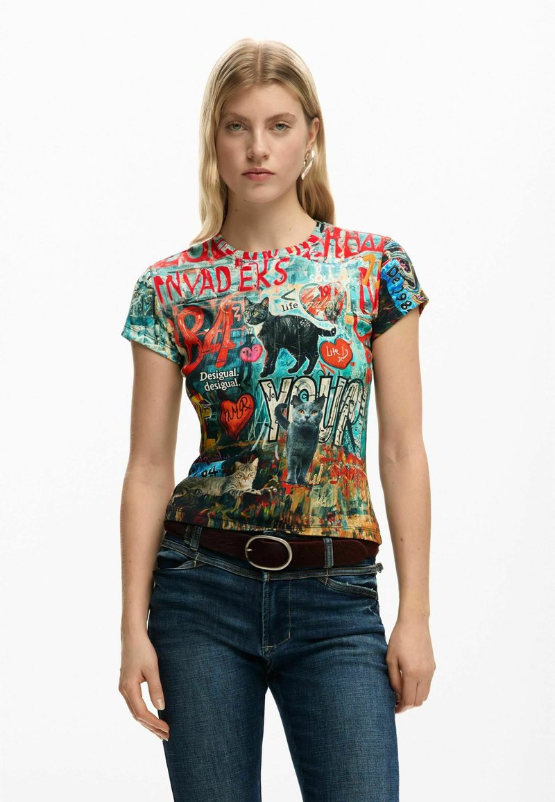 Graafinen t-paita, jossa on värikkäitä kissaprinttejä, numeroita ja tekstiä teksturoidulla kankaalla. Lyhyet hihat ja pyöreä kaula-aukko, yhdistetty tummiin farkkuihin.