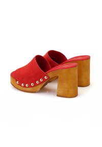 Rote Wildleder-Clogs mit einem breiten Riemen, verziert mit metallischen Nieten am Rand und einem klobigen Holzabsatz. Das Innenfutter hat eine kontrastierende rote Innensohle.