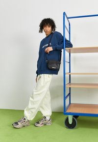 Modrá mikina s bílým textem, světle žluté volné kalhoty a šedé tenisky. Malá černá kabelka přes rameno. Dřevěný regál na kolečkách v pozadí.