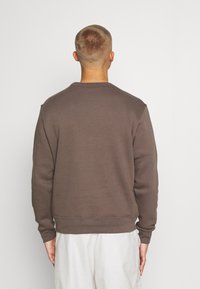 Muž s krátkými vlasy, oblečený ve hnědém dlouhém tričku a bílých kalhotách, stojí zády k jednoduchému pozadí.