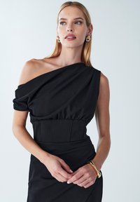 Abito nero con corpetto drappeggiato asimmetrico. Tessuto liscio, vita aderente con cuciture strutturate e un leggero lucido. Bracciale in oro come accessorio.