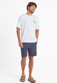 White cotton t-shirt with short sleeves and a small graphic print on the left chest, paired with navy blue shorts and brown sandals.
