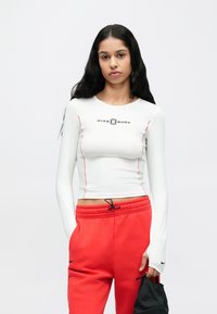 Bijela košulja s dugim rukavima s crvenim šavovima i logotipom Nike Shox, u kombinaciji s crvenim trenirkama i crnom torbom, s vrpcom za vezanje u struku.