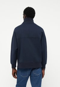 Navy blue sweatshirt with high collar, ribbed cuffs, and hem. Features a smooth texture and relaxed fit, paired with denim jeans.
