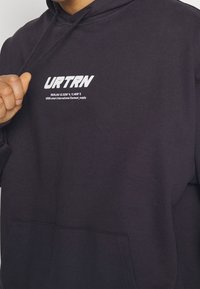 Persona con una sudadera oscura y el texto blanco "URTRN" y las coordenadas de Berlín en el pecho, sosteniendo un cordón cerca del cuello.