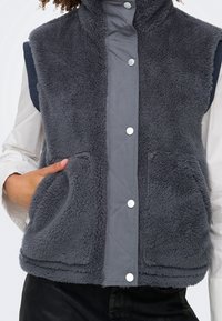 Personne portant un gilet polaire gris foncé avec des boutons-pression à l'avant, deux grandes poches plaquées et un col montant, sur une chemise blanche à manches longues.