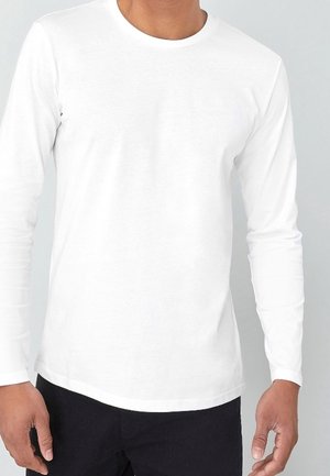 Homme portant un t-shirt blanc uni à manches longues avec col rond et un pantalon noir, debout devant un fond gris clair.