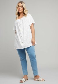 White linen blouse with a floral texture, round neckline, short sleeves, and wooden buttons. Paired with light blue jeans and white sandals.