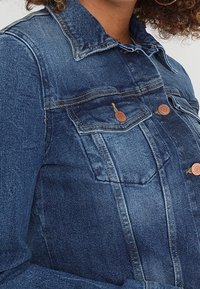 Džínová bunda v tmavě modré barvě s límcem, náprsními kapsami a knoflíky v měděném odstínu. Má klasický střih a mírně vybledlou texturu.