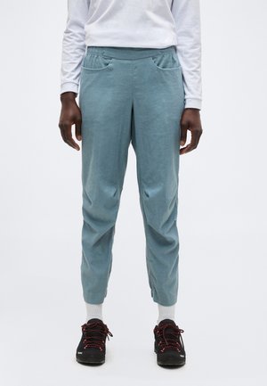 Dolná časť tela osoby, ktorá má na sebe svetlomodré teplákové nohavice, biele ponožky, čierne a červené tenisky a bielu košeľu s dlhým rukávom, stojaca pred jednofarebným pozadím.