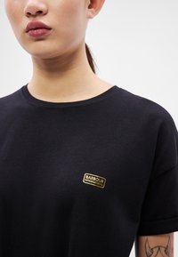 Μαύρο βαμβακερό μπλουζάκι με στρογγυλή λαιμόκοψη, με μικρό χρυσό ταμπελάκι λογότυπου "Barbour International" στο αριστερό μέρος του στήθους.