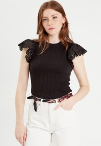 Top nero a coste con maniche a sbuffo in pizzo, abbinato a pantaloni corti bianchi a vita alta e una cintura a sciarpa con motivo.