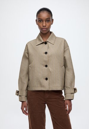 Un mannequin se tient devant un fond clair uni, vêtu d’une veste beige à carreaux avec des boutons noirs et d’un pantalon marron.