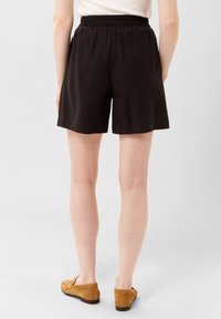 Černé šortky s elastickým pasem, rovný střih a měkká textura. V kombinaci s béžovými semišovými botami. Neutrální pozadí.