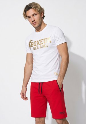 Giovane uomo con capelli biondi che indossa una maglietta bianca con testo dorato e pantaloncini rossi, in piedi davanti a uno sfondo bianco neutro.