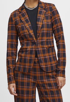 Mujer con blazer de cuadros marrón y azul marino y pantalones a juego sobre una camiseta negra con un collar de cadena plateada.