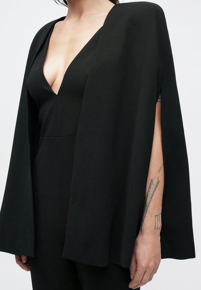 Mustast kangast keep langeb sügava V-kaelusega kombinesooveri peale, millel on avatud servad ja sile tekstuur. Käsivarrel on nähtav tätoveering.