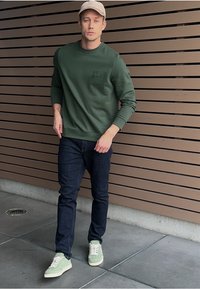 Zielona bluza, ciemne dżinsy, jasnooliwkowe buty, jasnobrązowa czapka. Bluza ma logo. Gładki materiał, dopasowany krój, styl casual.