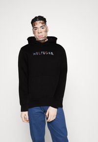 Svart hettegenser med en frontlomme og multikolorert "HILFIGER"-logo på brystet, sammen med blå bukser.