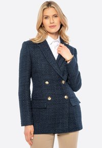 Dunkelblauer, doppelt knöpfbarer Blazer aus strukturiertem Stoff mit goldenen Knöpfen und zwei Fronttaschen. Über einem weißen Hemd gestylt.