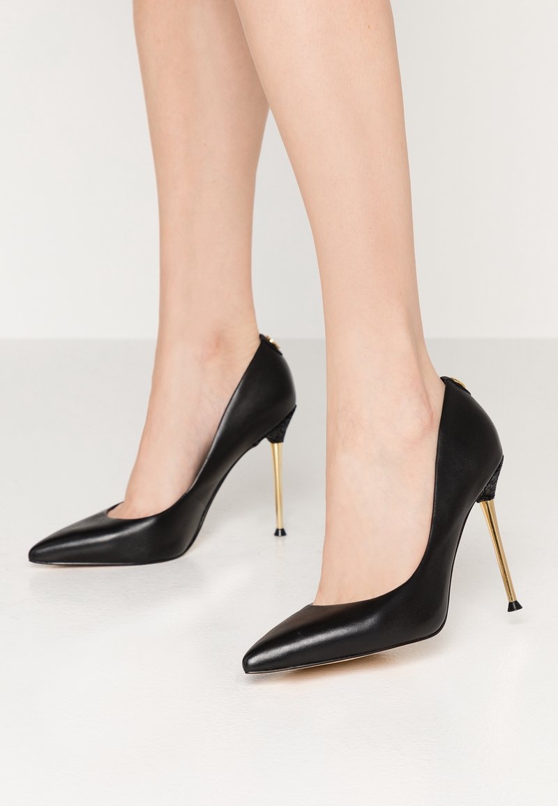 Escarpins noirs en cuir à bouts pointus avec talons aiguilles fins en or, portés pieds nus sur un fond uni.