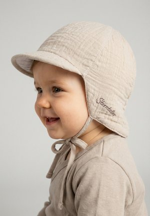 Smiling toddler wearing a beige quilted bonnet with chin ties and matching shirt, facing left on a neutral background.