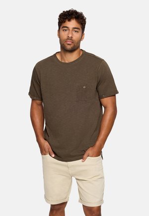 Hombre con camiseta de manga corta color verde oliva con bolsillo en el pecho y pantalones cortos beige remangados, de pie con las manos en los bolsillos.