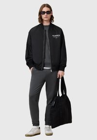 Chaqueta bomber negra con cremallera, sudadera gris, pantalones gris de corte slim, zapatillas blancas y una bolsa tote negra. Textura suave y diseño minimalista.