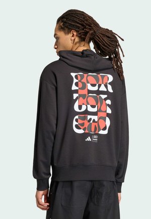 Osoba z dredami ubrana w czarną bluzę z kapturem z wyraźnym czerwono-białym abstrakcyjnym napisem oraz logo Adidas i Audi na plecach.